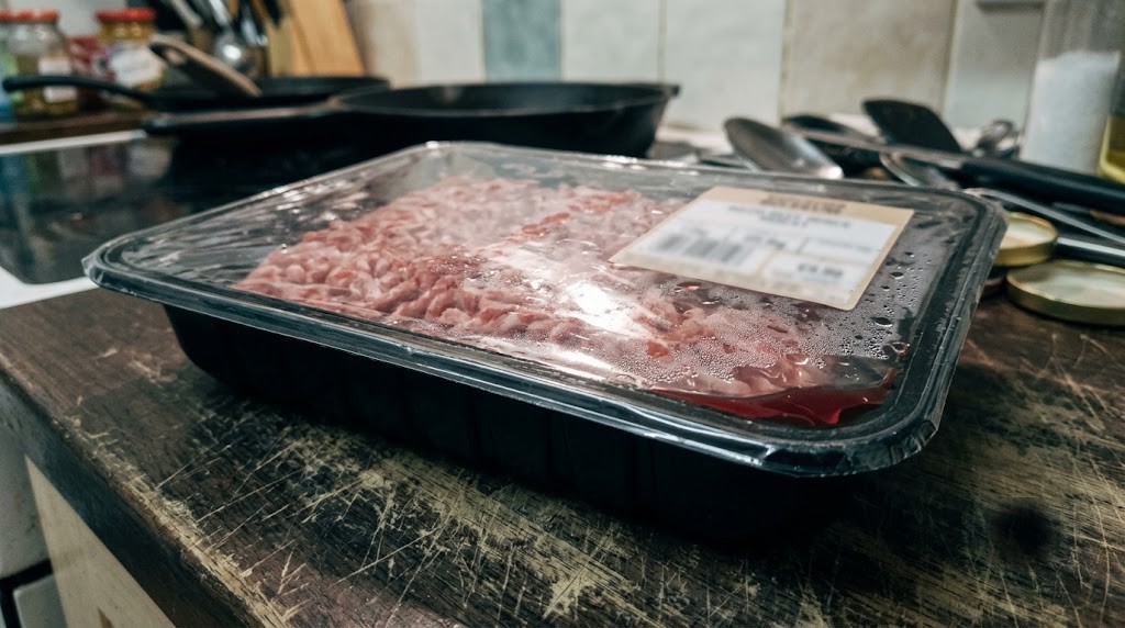 A supermarket pack of 80/20 ground beef mince on a scratched wooden kitchen worktop, cast-iron pans behind
